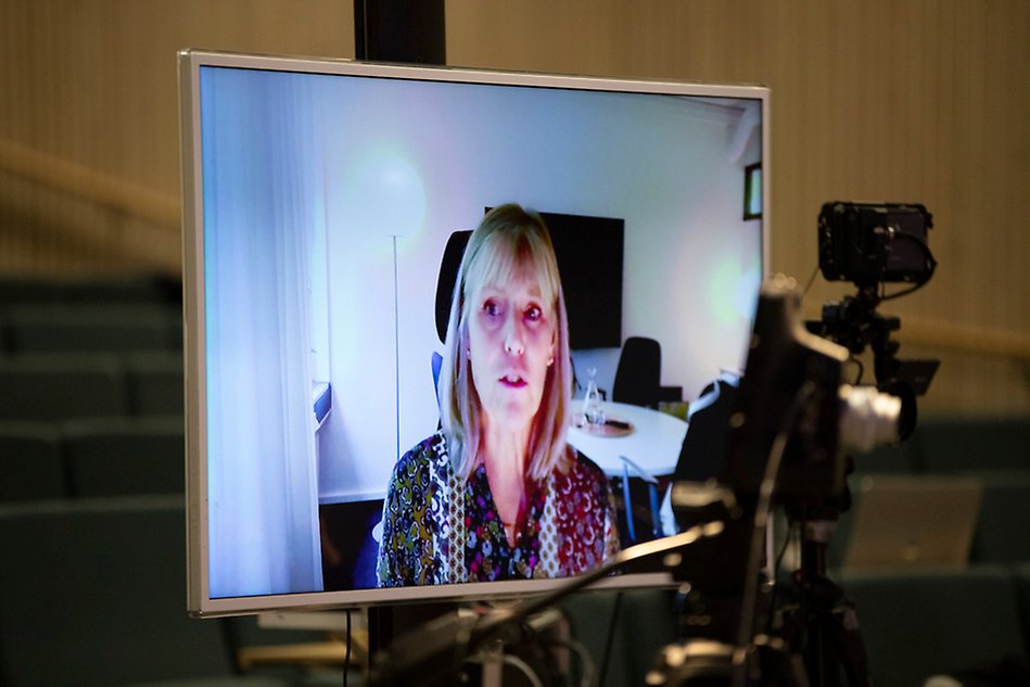 A woman is seen on a big screen talking. In the foreground there is a camera.