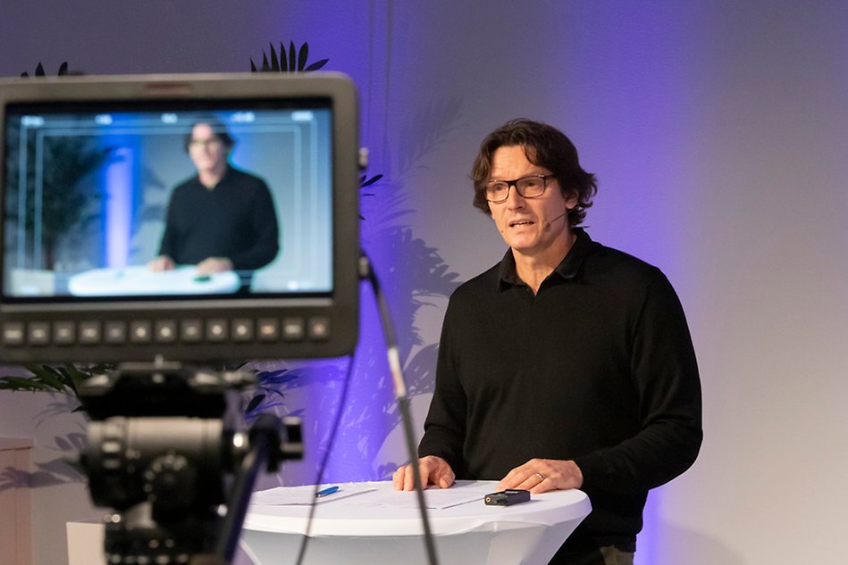 A man in dark clothes stands by a podium and looks into a camera. In the foreground a camera screen is seen. Photo.