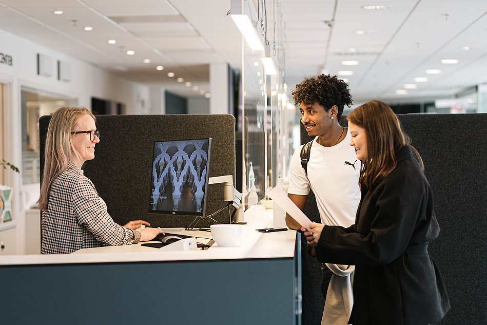 Två studenter talar med en kvinna i Servicecenters informationsdisk. Alla ler. Foto.