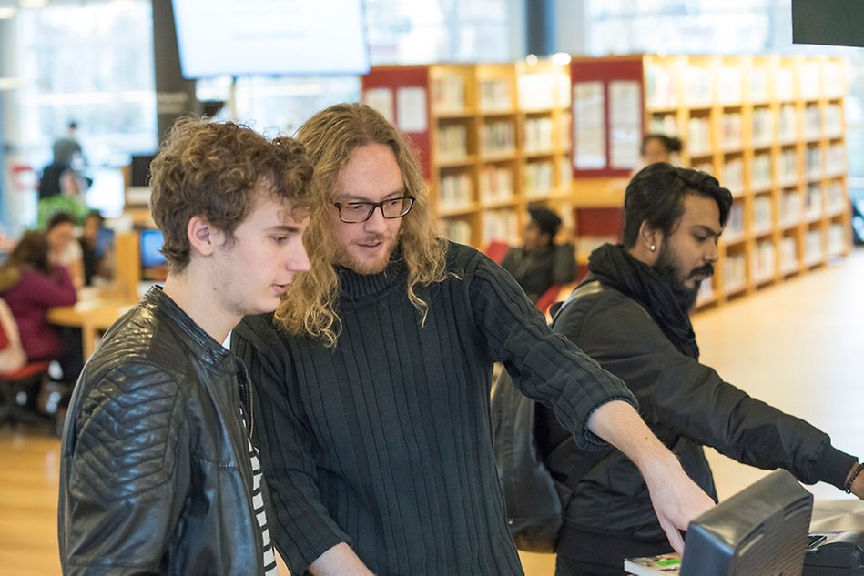 Man pekar och visar på en datorskärm för en annan man i biblioteksmiljö. Foto.