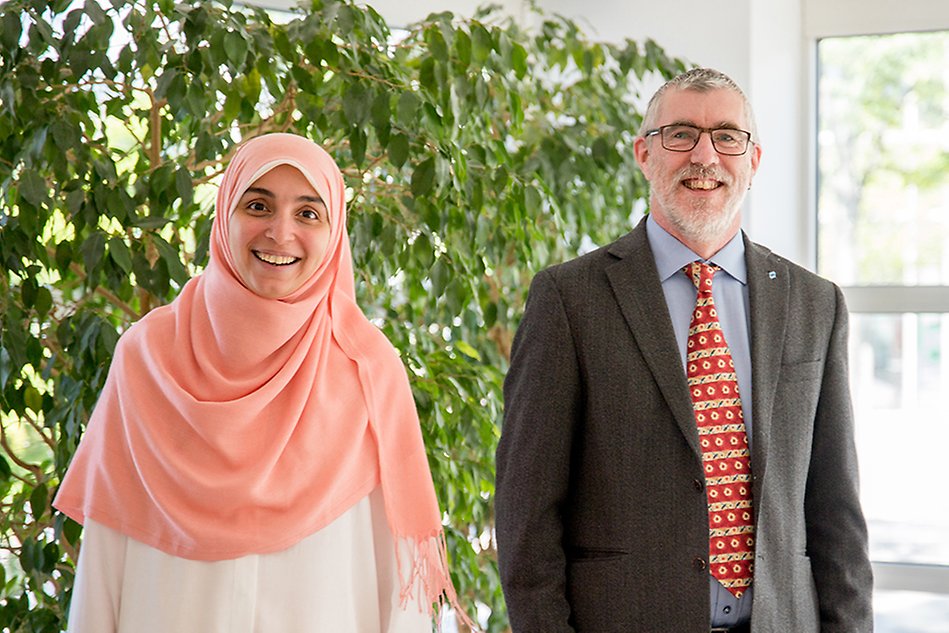 Woman wearing a pink hijab and a bearded man wearing a grey suit with a red tie, both smiling at the camera. Photo.