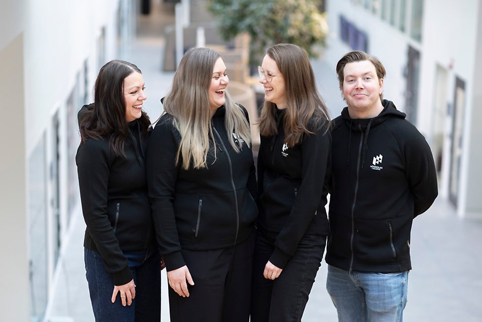 Four people are standing in a row in a bright environment indoors. They all have black shirts and are smiling and laughing. Photo.