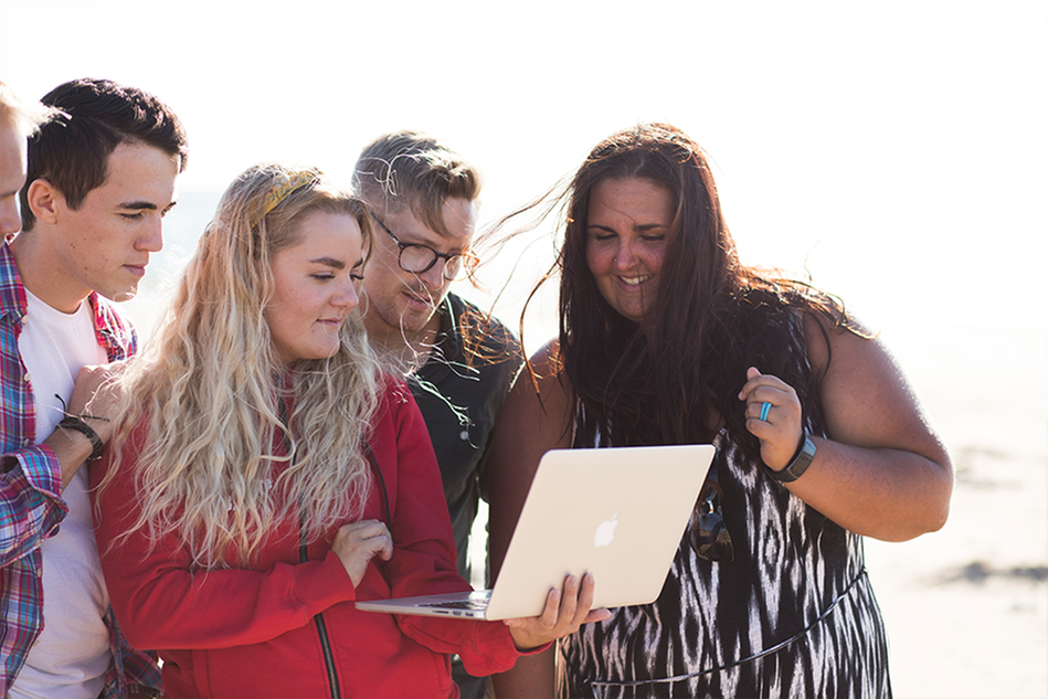 A group of students standing on the beach. One of them holding a laptop that everyone is looking at. Photo.