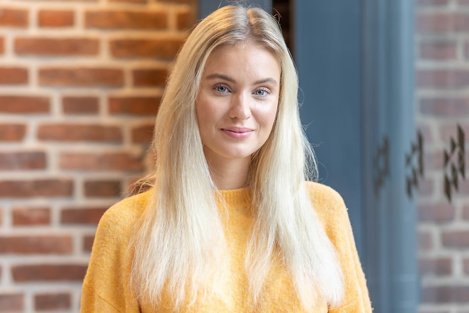 Porträtt på en ung kvinna med blont, utsläppt hår framför en tegelvägg. Foto.