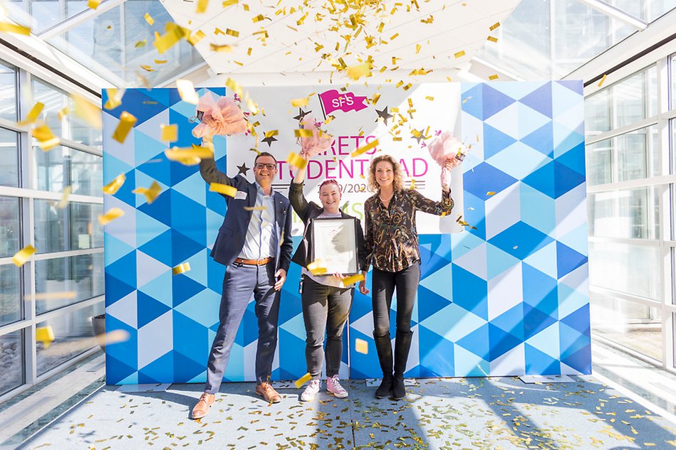 Three people on a stage holding diplomas and flowers with confetti raining down on them. Everyone smiling. Photo.