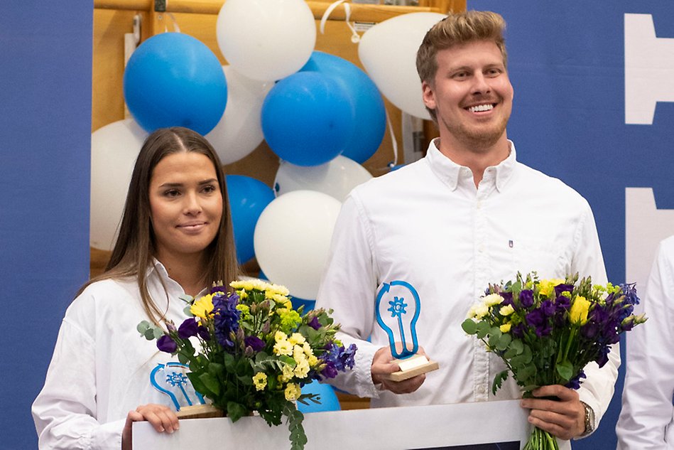 Two students in white shirts are holding bouquets of flowers, statuettes and a big check. Photo.