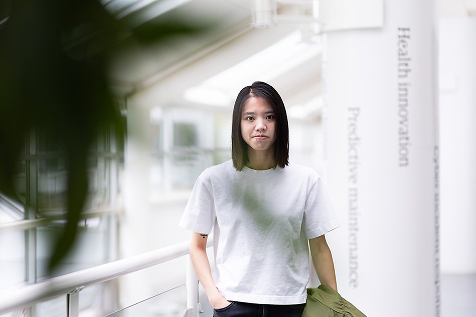Person with black hair looking into the camera. The background is a bright, airy space and a green plant can be seen in the foreground. Photo.
