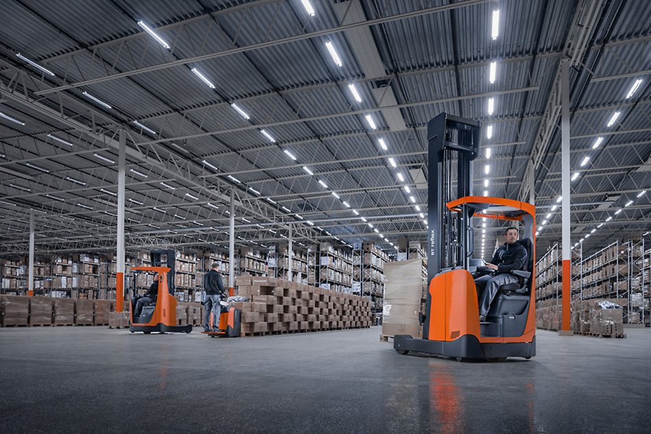 Two orange forklifts in a warehouse, one being operated by a person. Long shelves of packages can be seen in the background. Photo.