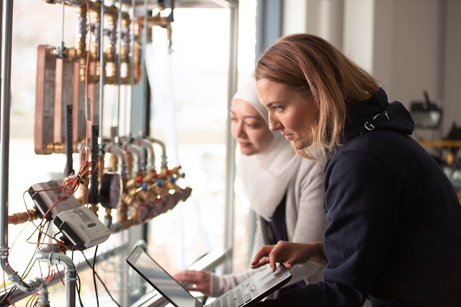 Two people looking at pipes and gauges. One is holding a laptop. Photo.