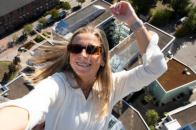 En person med solglasögon som ser ut att hänga i luften. Håret har blåst bakåt och under henne syns byggnader på marken. Foto.