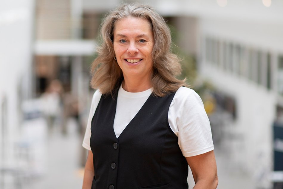 A person with long hair dressed in a white t-shirt and a black vest looking into the camera. Photo.