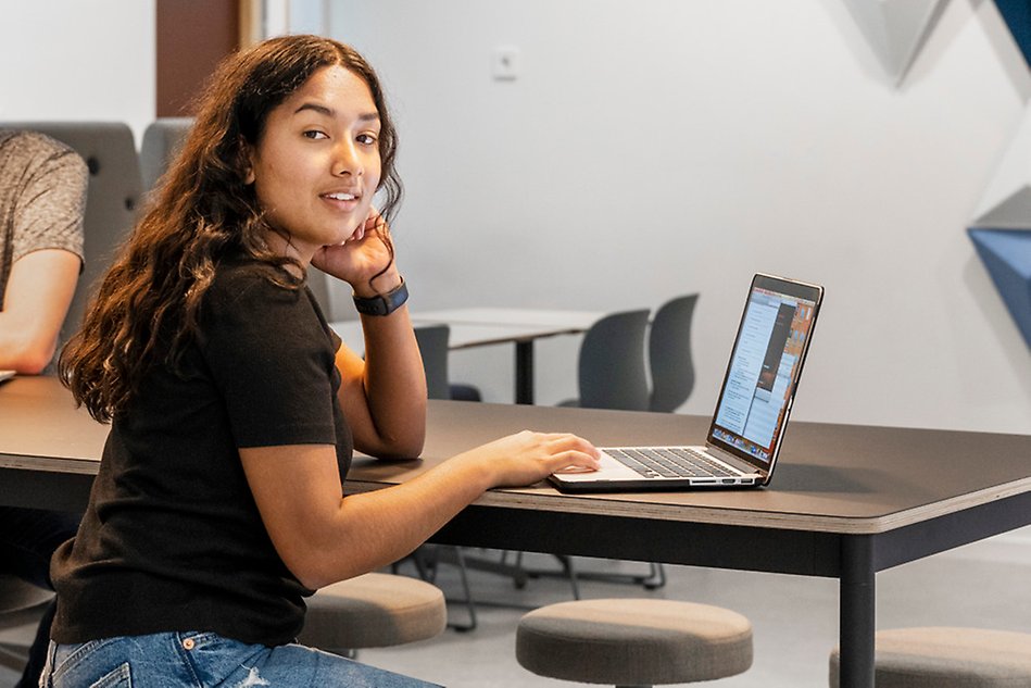 A younger women is sitting at a table with a laptop infront of her. The picture is taken from the side.  Photo.