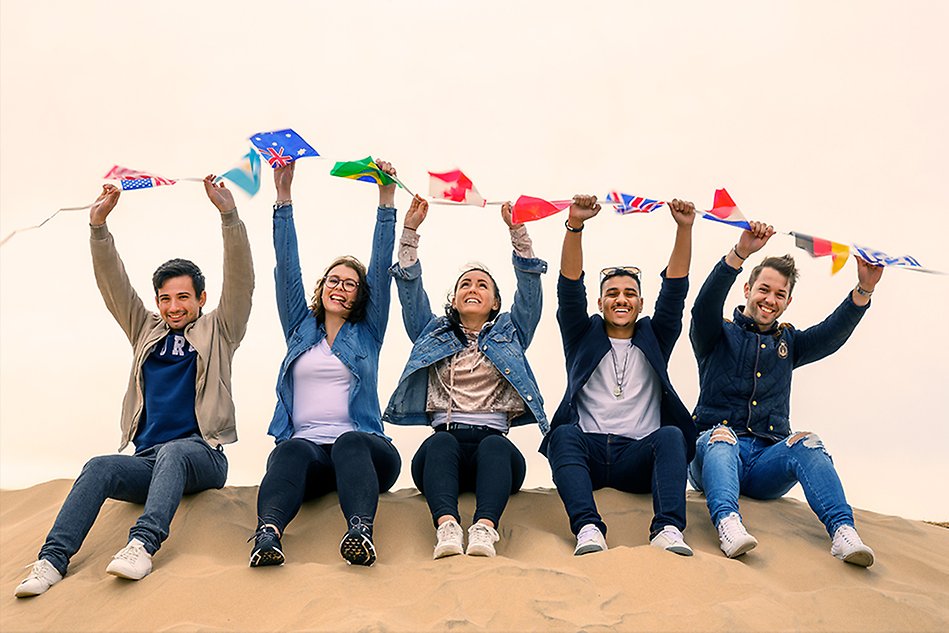 Five people sitting on a beach holding up a bunting with flags above their heads. Photo. 