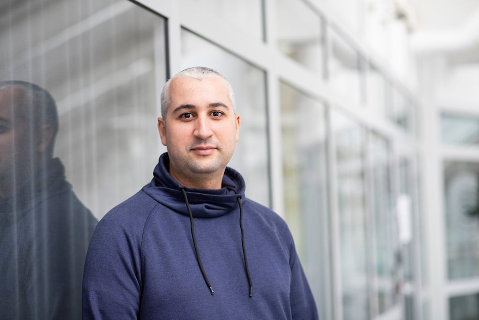 A man wearing a dark blue hoodie standing in front of a wall with glass window panes. Photo.