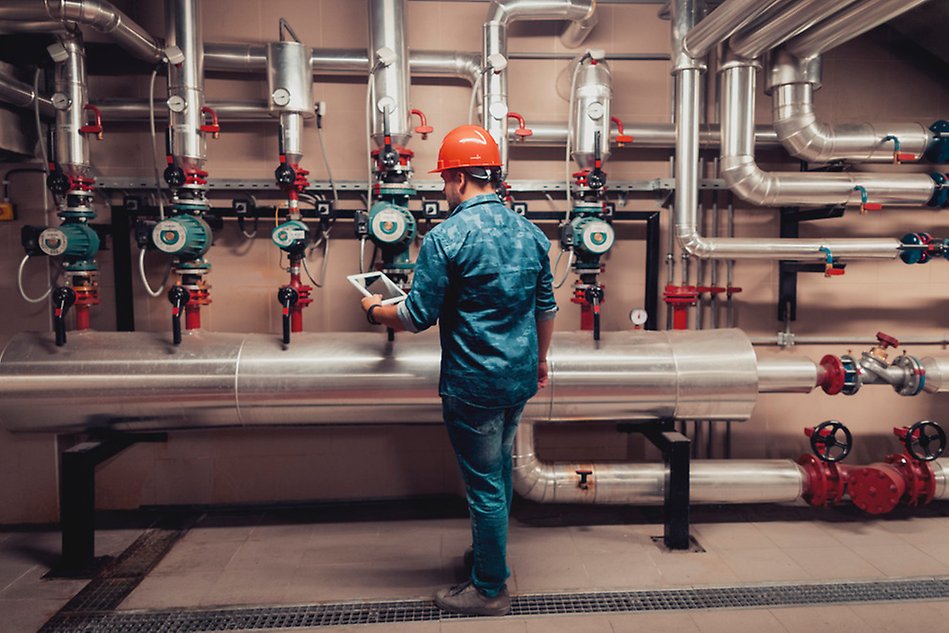 Man wearing a safety helmet standing with a padlet in front of a wall with pipes and valves. Photo.