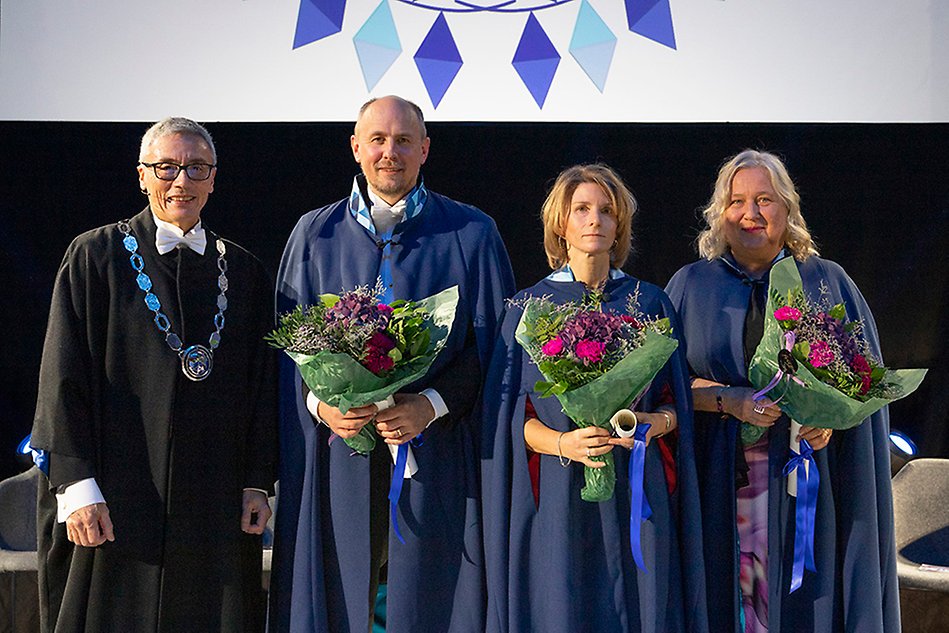 3 personer i blå caper står uppradade på en scen. I famnen har de blommor och diplom. Längst till vänster står en man i svart rektorskappa och med en stor silverkedja runt halsen Foto.