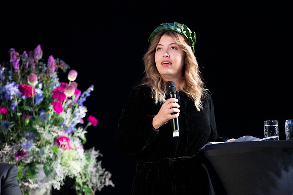 A person with long hair and a wreath on his head holding a microphone. Photography.