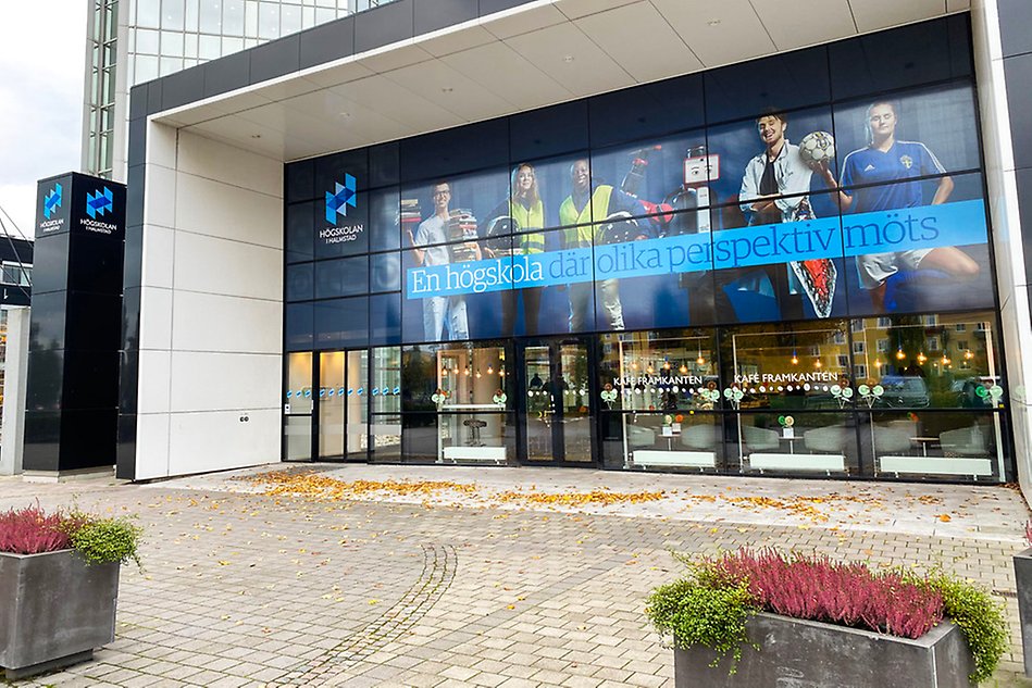 Högskolan i Halmstad entré med stor bild ovanför huvudingången på studenter och med texten En högskola där olika perspektiv möts. Växtlighet i förgrunden. Foto.