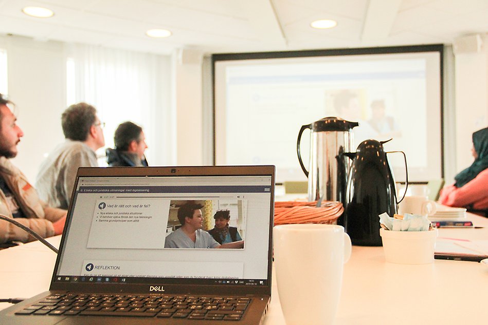 Bärbar dator på ett bord i ett konferensrum, flera människor sitter runt bordet och tittar på en digital skärm på väggen. Foto.