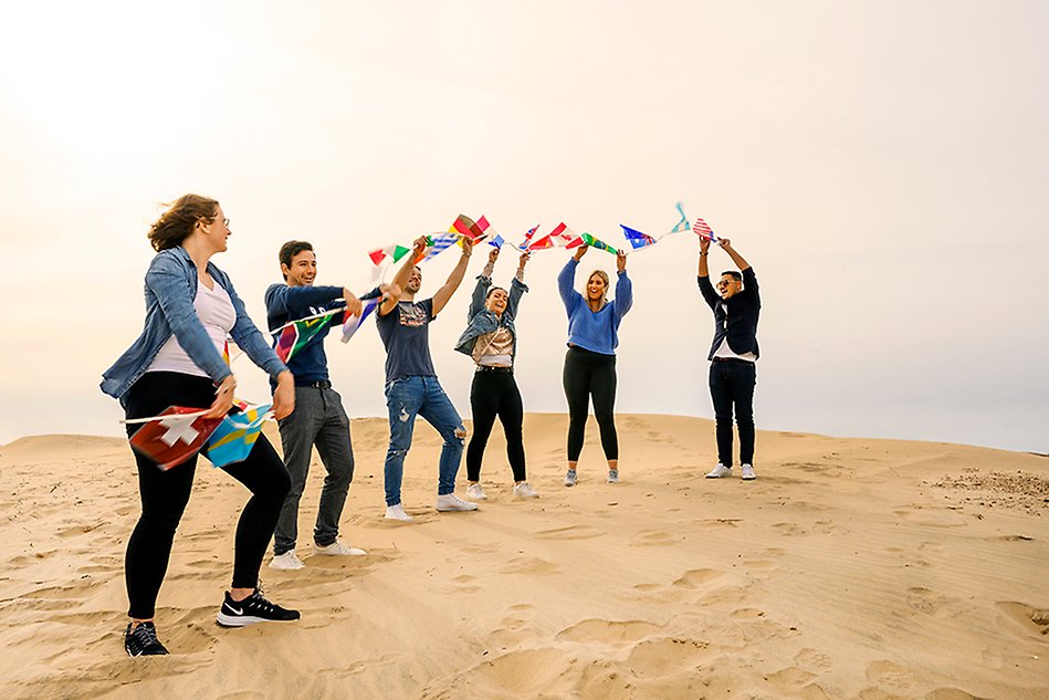 En rad med unga personer står på en strand och håller upp vimplar med flaggor från olika länder. Foto.