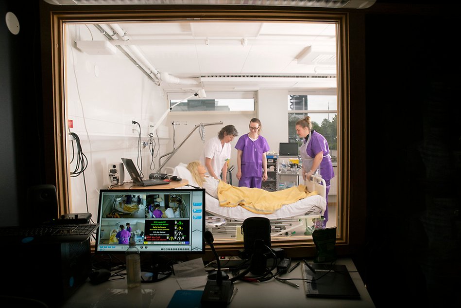 Three women in nursing clothes standing by a bed. The picture is taken from a observation room through a window. Photo.