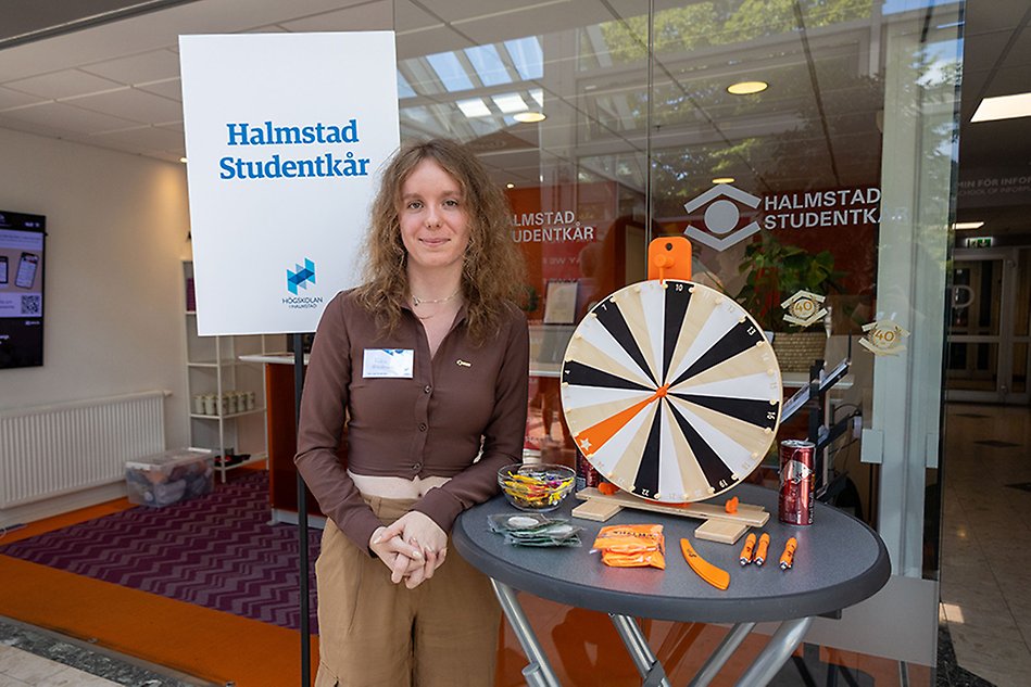 En ung person står vid ett bord med olika tävlingsprylar på. Personen ler och bakom syns texten "Halmstads Studentkår". Foto.